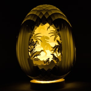 Może przedstawiać: Biała papierowa lampa w kształcie jajka z wyciętym wzorem dinozaura i palm w środku. Lampa jest oświetlona od wewnątrz, dając ciepłe żółte światło.