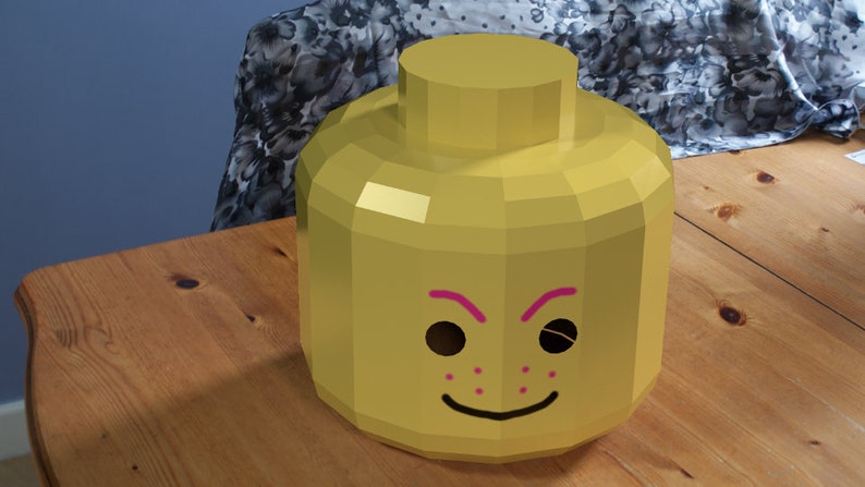 May include: A yellow Lego head costume with pink eyebrows and a smile. The head has black eyes and pink freckles.