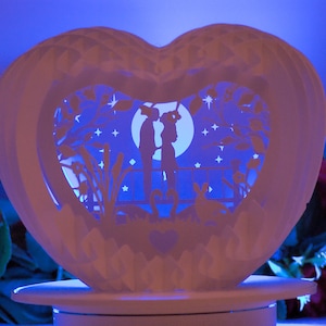 May include: A white, heart-shaped paper cut light with a romantic scene illuminated in blue. The scene features silhouettes of a couple, trees, stars, and a full moon. The light sits on a white base.