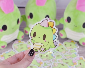 May include: A hand holds a green dinosaur sticker with a party hat and a heart-shaped detail. The sticker has a cartoon design with a light green body, a pink party hat, and a rainbow-colored spine. Several plush green dinosaurs with pink accents are in the background.