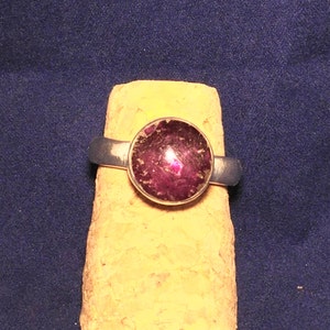 Può includere: Un anello d'argento con una pietra preziosa rotonda di colore viola scuro incastonata al centro.