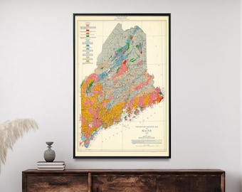 Op de afbeelding: Een vintage geologische kaart van Maine, ingelijst in zwart, hangt aan een witte muur boven een houten dressoir. De kaart is kleurgecodeerd om verschillende geologische formaties te tonen. De kaart heeft de titel "Geological Map of Maine".