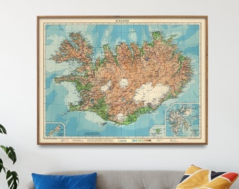 May include: A vintage map of Iceland in a wooden frame, hanging on a white wall above a light gray couch with blue and orange throw pillows. The map shows the island's geography and features a light blue ocean surrounding the island.
