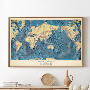 May include: A framed map titled "THE FLOOR OF THE OCEANS" shows a detailed, colourful relief of the ocean floor. The map is in a wooden frame and hangs on a white wall above a wooden cabinet. The map shows the world's oceans in shades of blue and the continents in shades of tan.