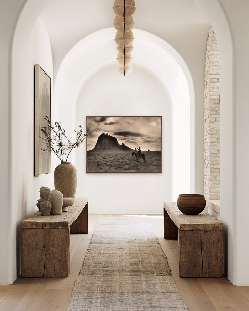May include: An interior hallway with a neutral color palette. The space features a framed sepia-toned landscape photograph, rustic wooden benches, and a woven runner. Decorative vases and a hanging light fixture add to the aesthetic.