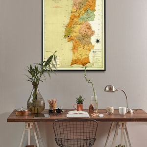 Puede incluir: Un mapa vintage de Portugal en un marco negro colgado en una pared gris claro sobre un escritorio de madera con una silla de alambre negro, un jarr&oacute;n con plantas verdes, una taza de cobre, una l&aacute;mpara y una taza blanca.