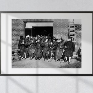 May include: A black and white photograph of a group of people in uniform, likely firefighters, sitting outside a fire station. They are all drinking from mugs or bottles.