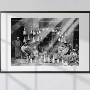 May include: Black and white photo of a group of men working in a shop. The men are sitting and standing around a table filled with copper and brass pots and jugs. The shop is lit by sunlight streaming through the windows.