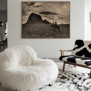 May include: A sepia-toned framed print of a person on horseback in a desert landscape with a large mountain in the background. A white, fluffy, round chair and a cowhide-patterned chair sit on a white and black rug.