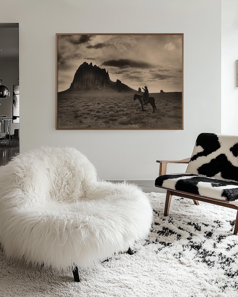 May include: A sepia-toned framed print of a person on horseback in a desert landscape with a large mountain in the background. A white, fluffy, round chair and a cowhide-patterned chair sit on a white and black rug.