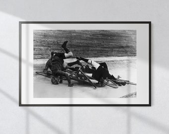 May include: A black and white photograph of two cyclists who have crashed on a track. One cyclist is lying on their back, while the other is upside down with their legs in the air.