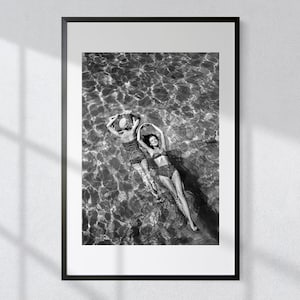 May include: Black and white photograph of two people floating in a pool of water. The people are wearing swimsuits and are looking up at the camera.