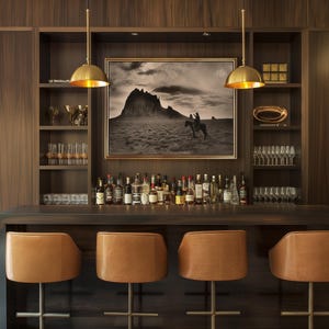May include: A home bar with a dark wood finish, featuring a collection of liquor bottles, glassware, and leather bar stools. A framed black and white landscape painting hangs above the bar. Two gold pendant lights illuminate the bar area.