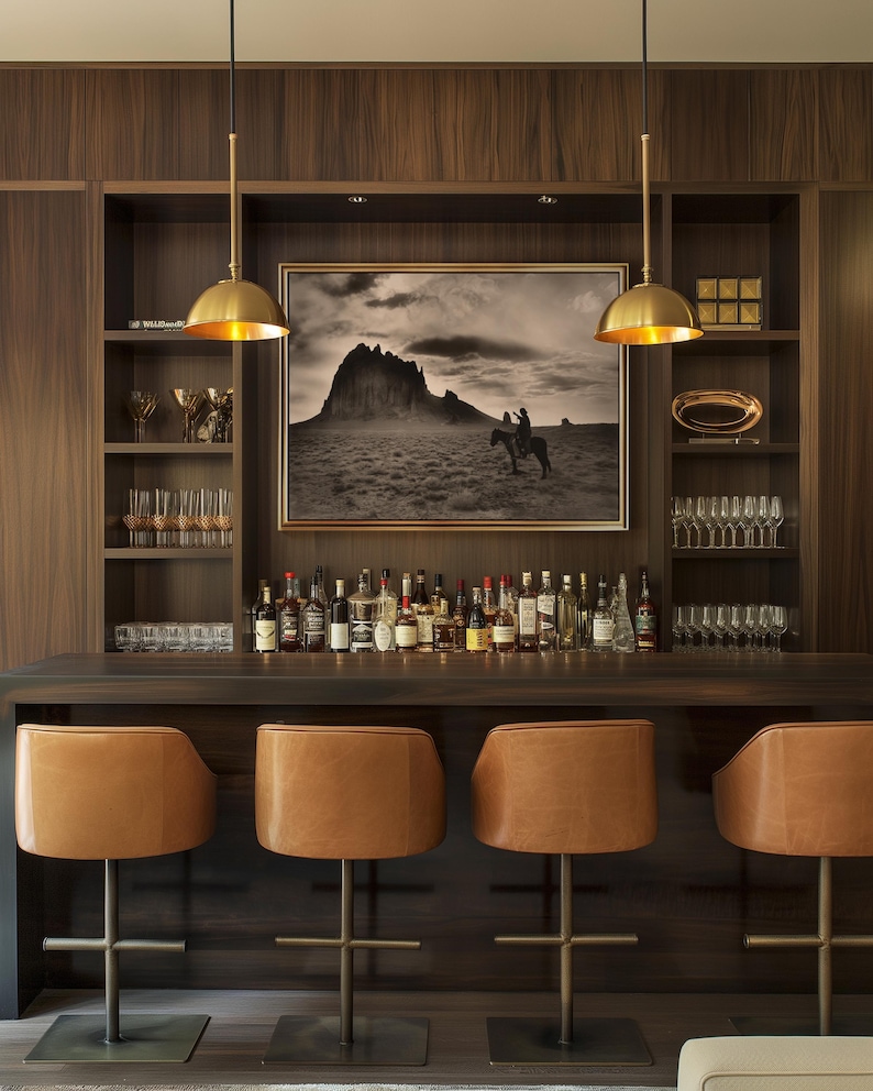 May include: A home bar with a dark wood finish, featuring a collection of liquor bottles, glassware, and leather bar stools. A framed black and white landscape painting hangs above the bar. Two gold pendant lights illuminate the bar area.