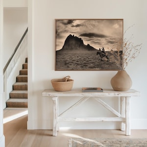 May include: A sepia-toned framed print of a mountain landscape hangs above a rustic white wooden console table. A woven basket, a small book, and a ceramic vase with dried branches are on the table. A staircase is visible to the left.