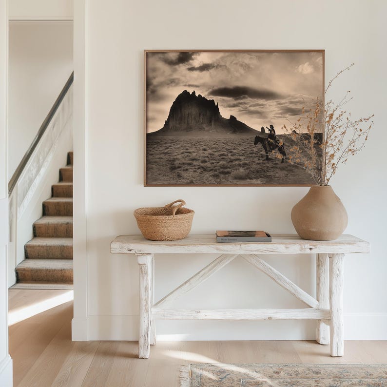 May include: A sepia-toned framed print of a mountain landscape hangs above a rustic white wooden console table. A woven basket, a small book, and a ceramic vase with dried branches are on the table. A staircase is visible to the left.