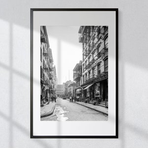May include: A black and white photograph of a narrow street in a city. The street is lined with tall buildings, and there are people walking on the pavement. The photo is framed in a black frame.
