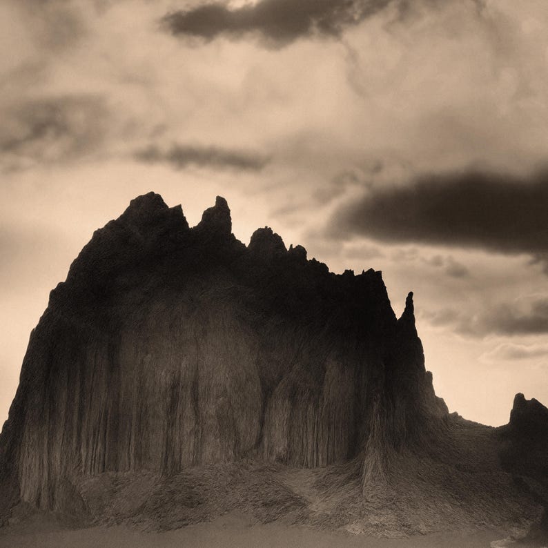 May include: Sepia-toned photograph of a mountain range under a cloudy sky. The mountain has a rugged, textured surface with vertical striations. The sky is filled with dark, ominous clouds, creating a dramatic and moody landscape.