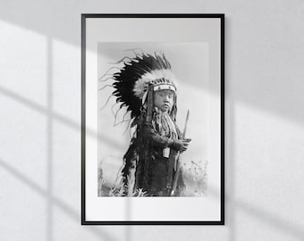 May include: Black and white photograph of a young person wearing a traditional Native American headdress and holding a spear. The person is standing in a field with tall grass.
