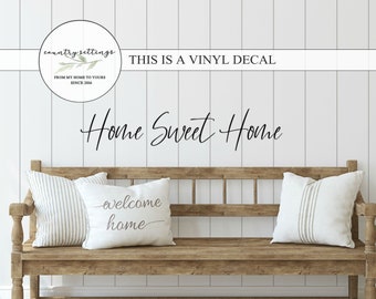 May include: A wooden bench with two pillows, one with the text "welcome home" and the other with a striped pattern. The wall behind the bench has a white vinyl decal that reads "Home Sweet Home" and a logo that says "country settings from my home to yours since 2006".
