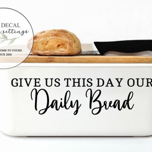 May include: White bread box with a bamboo lid, a loaf of bread, and a black knife. The box has the text "GIVE US THIS DAY OUR Daily Bread". The image also includes the text "VINYL DECAL country settings FROM MY HOME TO YOURS SINCE 2016".