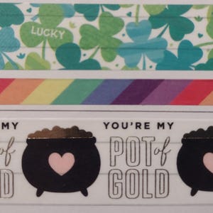 May include: A white and green patterned washi tape with the word "Lucky" printed on it. Below the tape is a rainbow patterned washi tape. The bottom tape is white with the text "You're my pot of gold" and a black cauldron with a pink heart.
