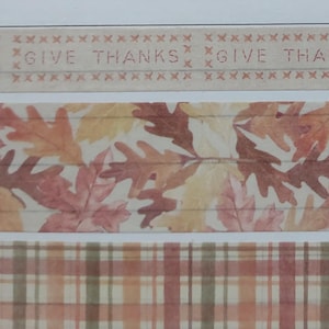 May include: Decorative paper tape rolls with autumn themes. The top roll has the words "GIVE THANKS" in a rustic font. The middle roll features fall leaves in shades of orange, red, and yellow. The bottom roll has a plaid pattern in similar colors.