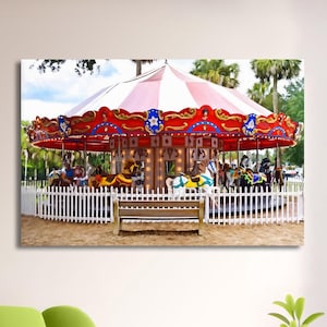 May include: A wall art print featuring a carousel with a red and white striped canopy, ornate red trim with blue star accents, and various painted horses. A wooden bench sits in front of a white picket fence surrounding the ride.