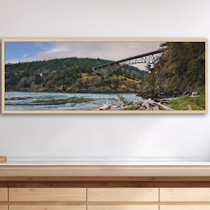 May include: A framed landscape painting depicts a bridge spanning a body of water, with forested hills in the background. The artwork features a natural wood frame and captures the scenic beauty of the area.