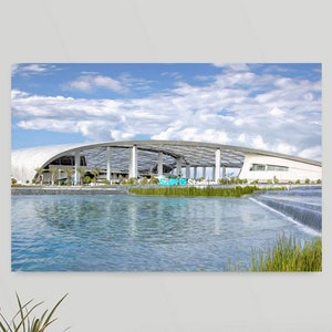 May include: A scenic canvas print of SoFi Stadium, a modern architectural marvel. The image captures the stadium's unique design, with a curved roof and open-air structure. A serene water feature and blue sky with clouds complete the scene.