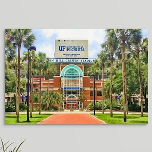 May include: A canvas print of Ben Hill Griffin Stadium at the University of Florida. The stadium is a large brick building with a blue and white sign at the top. Palm trees line the sides of the stadium, and a red brick path leads to the entrance.