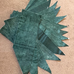 May include: A stack of ten green fabric triangles, each with a slightly different shade of green. The fabric has a textured, almost speckled appearance.