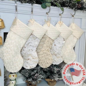 Peut inclure: Cinq bas de Noël crème avec des dessus matelassés et des corps texturés. Chaque bas est orné d'un ornement en bois. Ils sont suspendus à une cheminée blanche, créant une ambiance festive pour les fêtes.