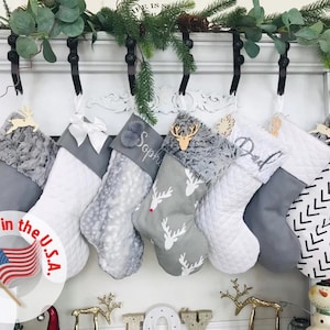May include: Six gray and white Christmas stockings with various designs, including reindeer, snowflakes, and geometric patterns, hanging on hooks above a fireplace. The stockings are personalized with names like "Sophia" and "Dad".