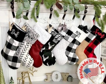Puede incluir: Conjunto de seis calcetines de Navidad personalizados con tela de cuadros negros y blancos. Los calcetines están colgados en una repisa de chimenea blanca y llevan los nombres "Papá", "Mamá", "Sam", "Ella" y otros dos. Los calcetines están hechos a mano en los Estados Unidos.