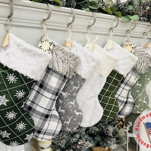 May include: A collection of Christmas stockings with various patterns, including snowflakes, plaid, and deer. Each stocking has a white faux fur cuff and a wooden ornament. The stockings are hanging on a white mantelpiece.
