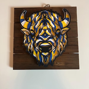 以下が含まれることがあります： 青、黄色、白の色の重ねられた紙切りデザインで、バッファローの頭が描かれた木製の壁飾りです。
