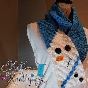 May include: A white crocheted snowman scarf with blue accents. The scarf has a blue crocheted border and a blue crocheted nose. The scarf is on a white mannequin.