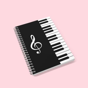 Puede incluir: Cuaderno con espiral con una cubierta negra con una clave de sol blanca y un diseño de teclado de piano. Las páginas del cuaderno se mantienen unidas por una encuadernación en espiral plateada. El diseño del teclado está en el lado derecho.