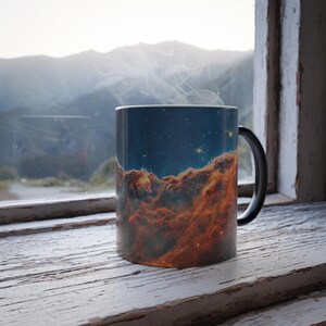 May include: A black coffee mug with a colorful nebula design. The mug is filled with a steaming beverage and is sitting on a wooden surface near a window.
