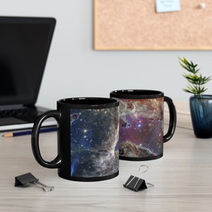 May include: Two black ceramic mugs with a space nebula design. The mugs have a black rim and handle. The nebula design is a colorful mix of blue, purple, pink, and white.