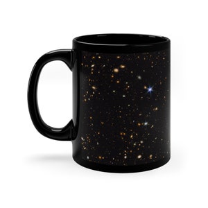 Puede incluir: Taza de café de cerámica negra con una imagen a todo color de un cielo nocturno estrellado. La imagen muestra una variedad de estrellas y galaxias en un esquema de color azul oscuro y negro.