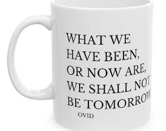 Puede incluir: Taza de cerámica blanca con asa curva. La taza presenta texto negro que dice "WHAT WE HAVE BEEN, OR NOW ARE, WE SHALL NOT BE TOMORROW" con "OVID" debajo. La taza es de tamaño estándar.