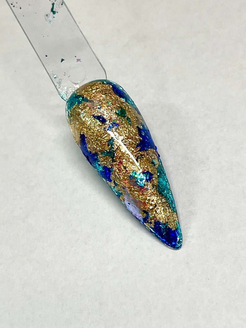 May include: A close-up of a long, pointed, blue and gold nail with a metallic finish. The nail is decorated with gold flakes and blue and green glitter.