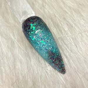 May include: A nail swatch with a gradient design. The top is dark teal, fading to a lighter teal with iridescent glitter. The glitter has teal, purple, and silver flakes, creating a shimmer. The swatch is on a clear stick.