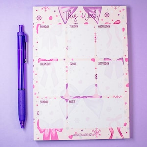 Può includere: Una penna viola è appoggiata accanto a un'agenda settimanale. L'agenda presenta uno sfondo bianco con illustrazioni di fiocchi rosa e viola, fiocchi di neve e la scritta "This Week". I giorni della settimana sono etichettati, con spazio per le note.