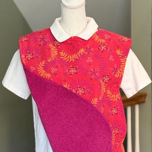 May include: A white collared shirt with a decorative bib. The bib features a vibrant floral pattern in shades of pink, orange, and yellow. A solid magenta fabric is layered over the floral pattern, creating a unique design.