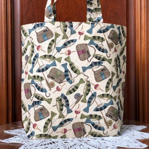 May include: A beige tote bag with a repeating pattern of fish, tackle boxes, and bobbers. The bag has two handles and is made of fabric. The background is a wooden door.