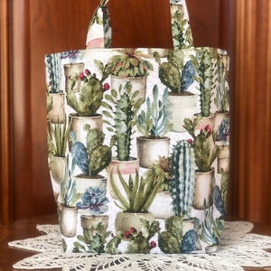 May include: A white tote bag with a green and brown cactus print. The bag has a single handle and is made of fabric.