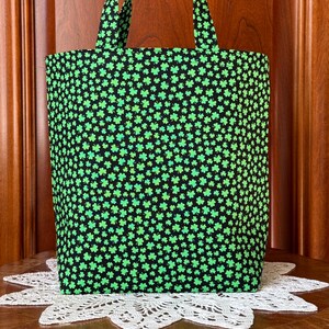 May include: A black tote bag with a repeating pattern of green shamrocks. The bag has two handles and is made of fabric. The shamrocks are in various shades of green, creating a festive look. The bag is sitting on a white lace doily.